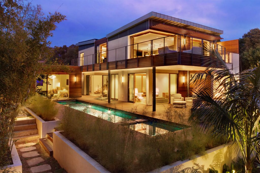 A modern two-storey beach house at dusk with a lap pool, timber cladding and full-height glazing.
